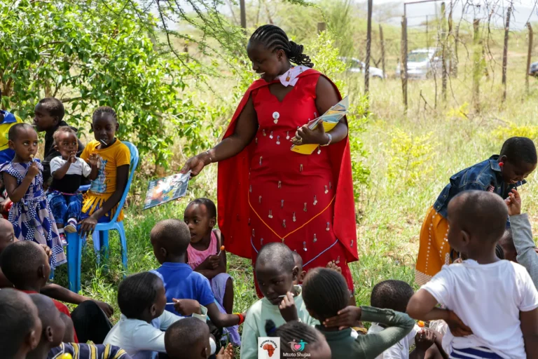 A woman in a red shuka speaks to children seated on the grass at an outdoor reading session in Kenya