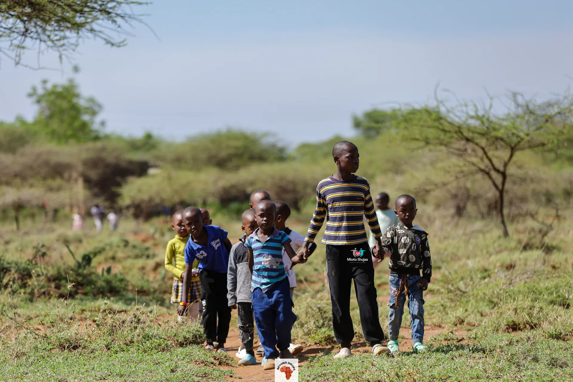 Children walk along a dirt path through dry savanna with acacia bushes in rural Kenya