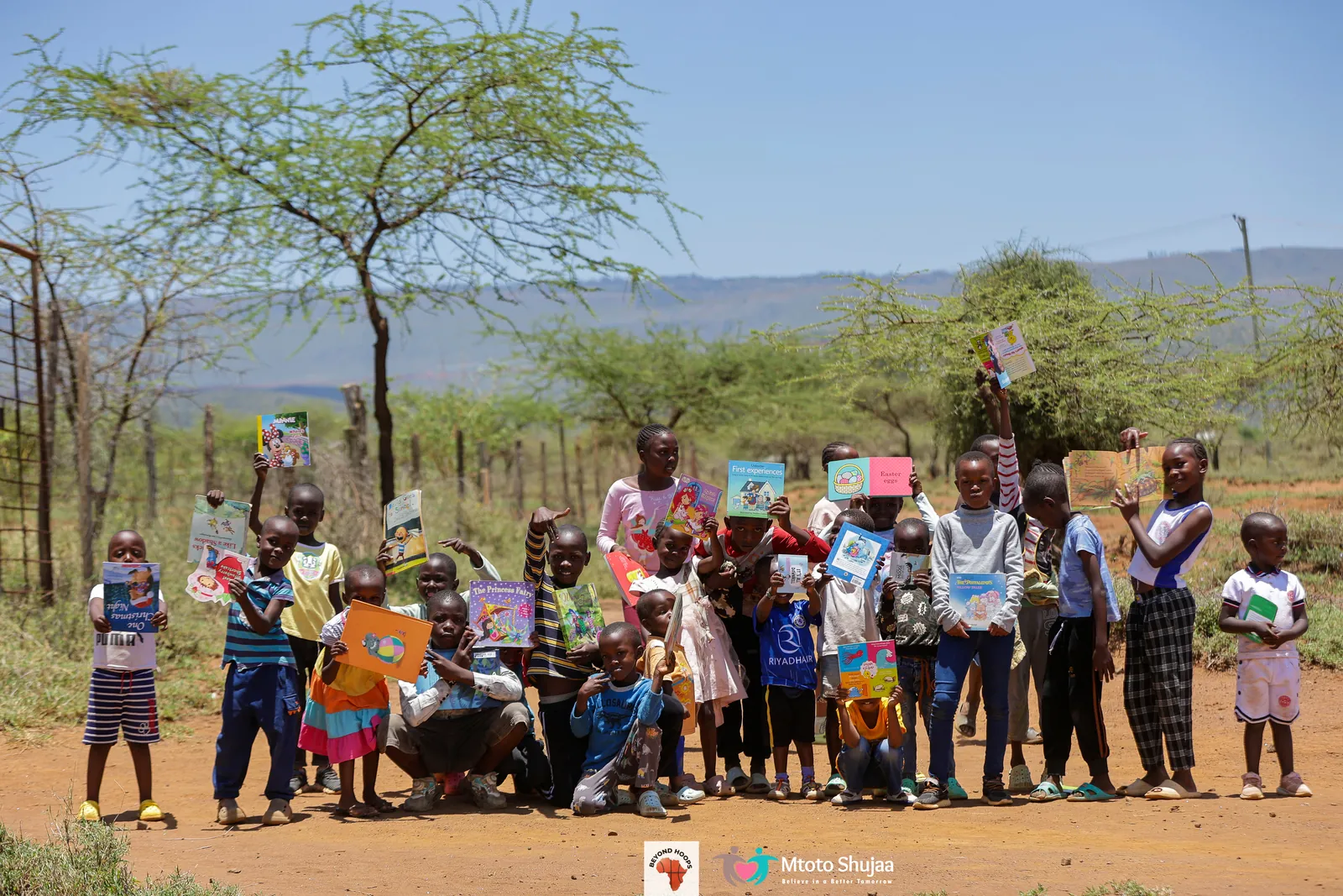 Children and volunteers hold up books in open savanna dotted with acacia trees in rural Kenya