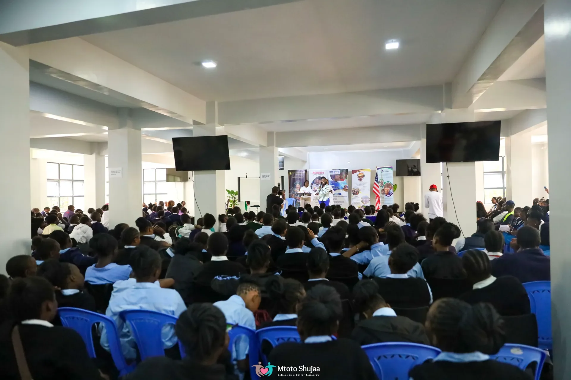 A large audience sits in a bright hall facing screens during a Mtoto Shujaa community forum in Kenya