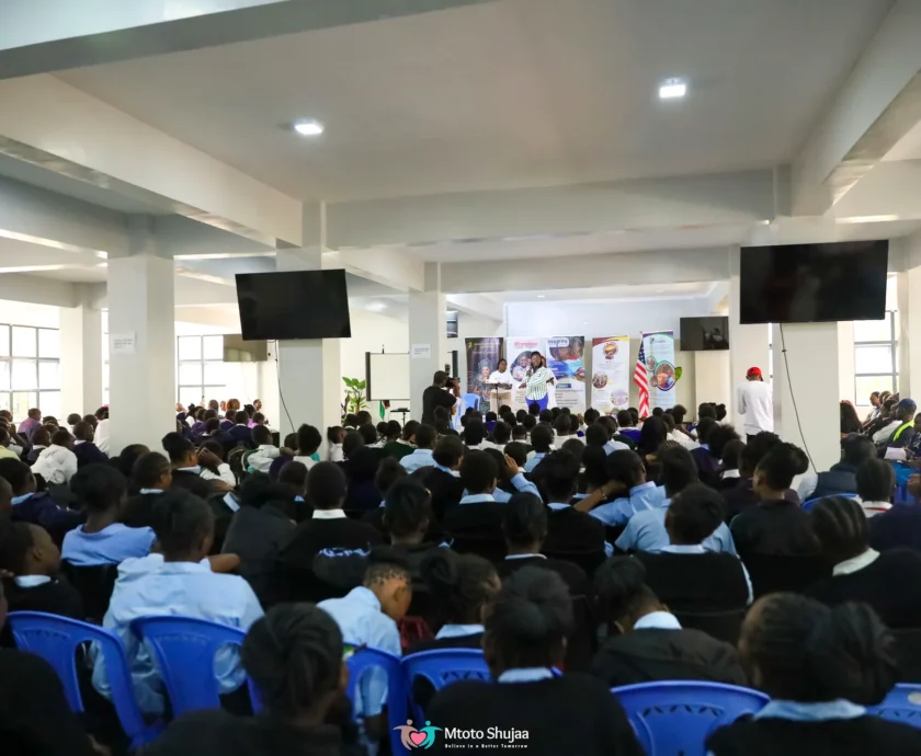 A large audience sits in a bright hall facing screens during a Mtoto Shujaa community forum in Kenya