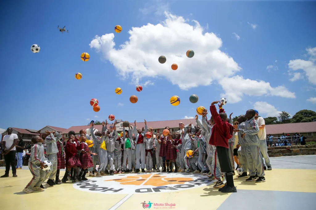 Children and coaches throw basketballs high into a blue sky on an outdoor court in Kenya
