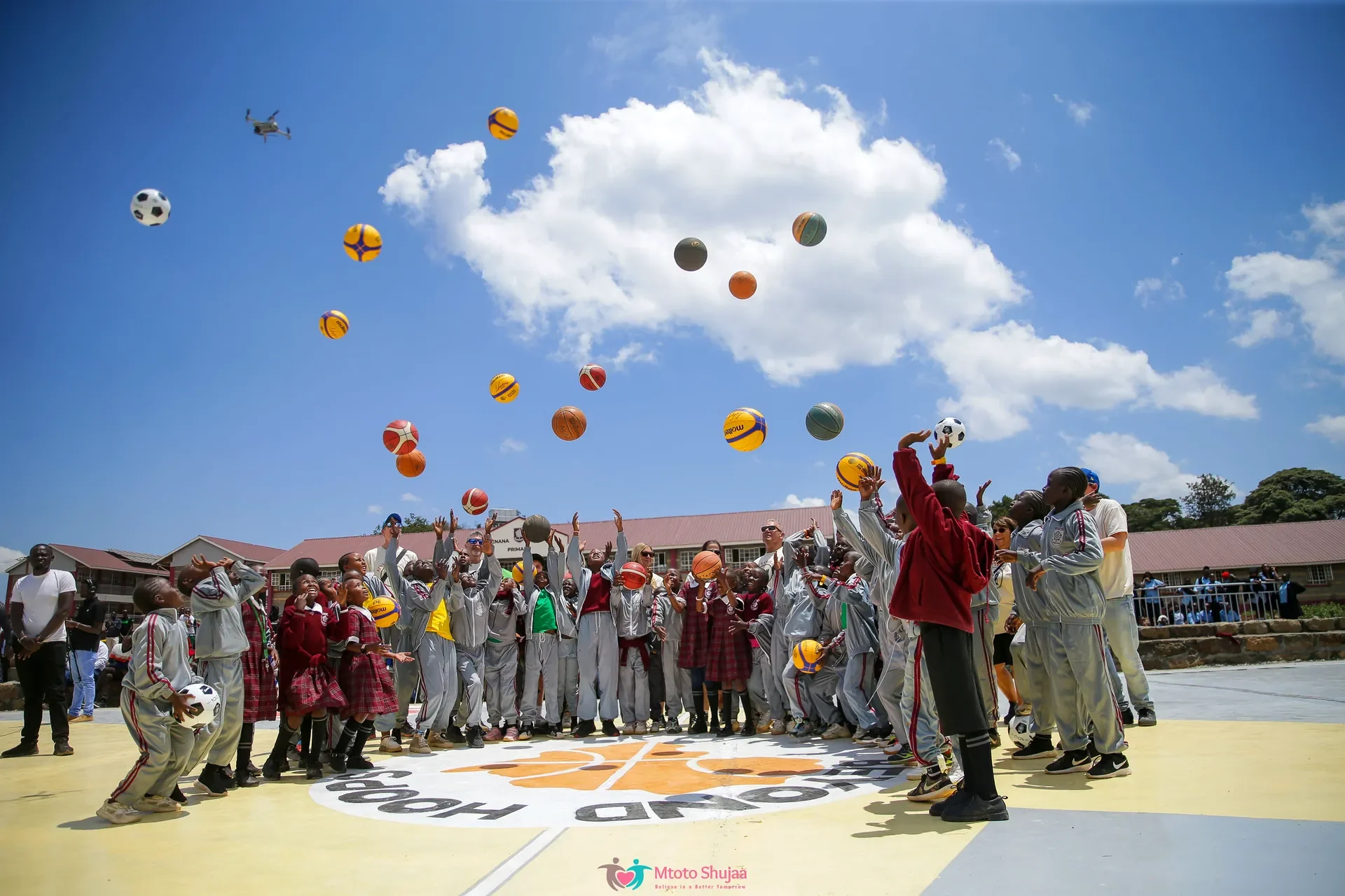 Children and coaches throw basketballs high into a blue sky on an outdoor court in Kenya