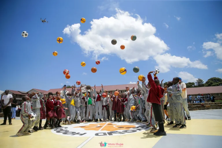 Children and coaches throw basketballs high into a blue sky on an outdoor court in Kenya