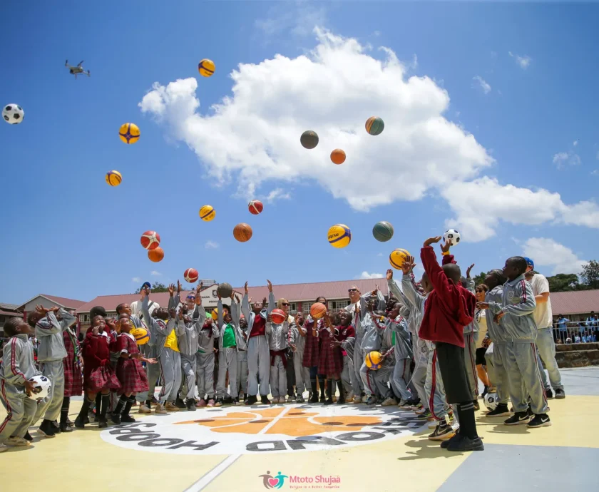 Children and coaches throw basketballs high into a blue sky on an outdoor court in Kenya