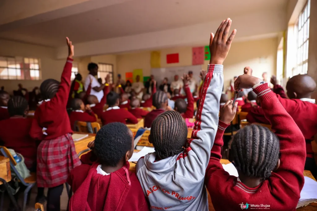 Students in red school uniforms raise their hands eagerly in a school hall in Kenya