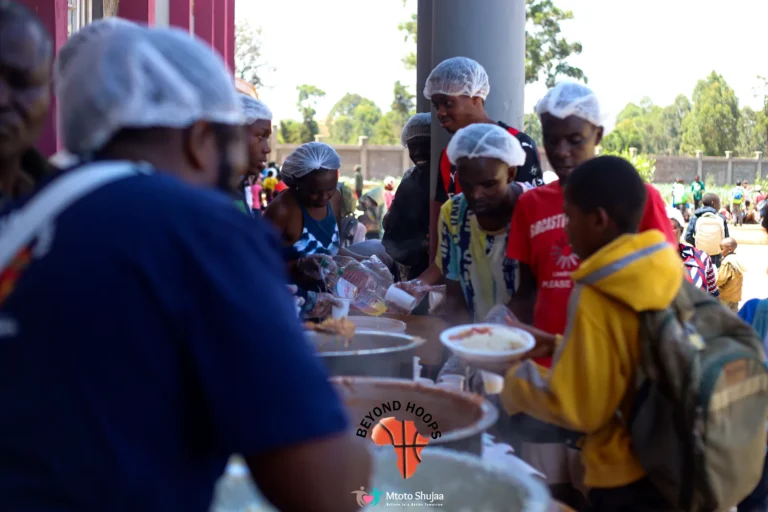 Volunteers wearing hairnets serve food to children at an outdoor feeding day in Kenya