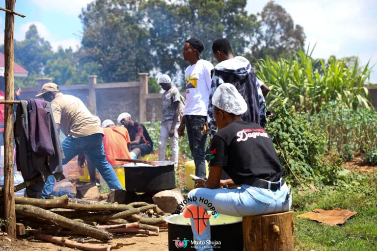 A cook prepares food in a large pot over a firewood stove outdoors in Kenya